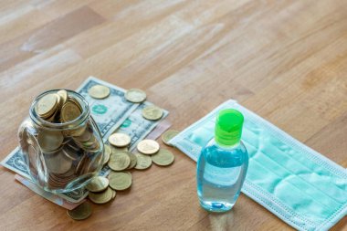 Masadaki madeni paralar ve yüz maskesi ve el temizleme jeli gibi dolar banknotları. Corona virüsü salgını sırasında pahalı yüz maskesi ve fotokopi alanı ile el jeli..
