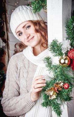 Beyaz örgü şapkalı, kırmızı saçlı bir kız ve örgü süveter Noel tarzında dekore edilmiş bir verandada duruyor. Kış