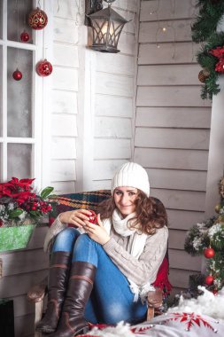 Beyaz örgü şapkalı, kırmızı saçlı bir kız ve örgü bir süveter Noel ağacının yanındaki verandada Noel tarzında süslenmiş bir sandalyede oturuyor. Kış