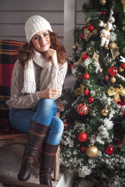 Beyaz örgü şapkalı, kırmızı saçlı bir kız ve örgü bir süveter Noel ağacının yanındaki verandada Noel tarzında süslenmiş bir sandalyede oturuyor. Kış