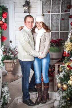 Beyaz örgü süveter giyen bir adam ve bir kız Noel tarzında dekore edilmiş bir verandada oturuyorlar. Kış