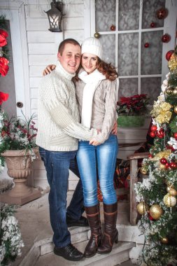 Beyaz örgü süveter giyen bir adam ve bir kız Noel tarzında dekore edilmiş bir verandada oturuyorlar. Kış