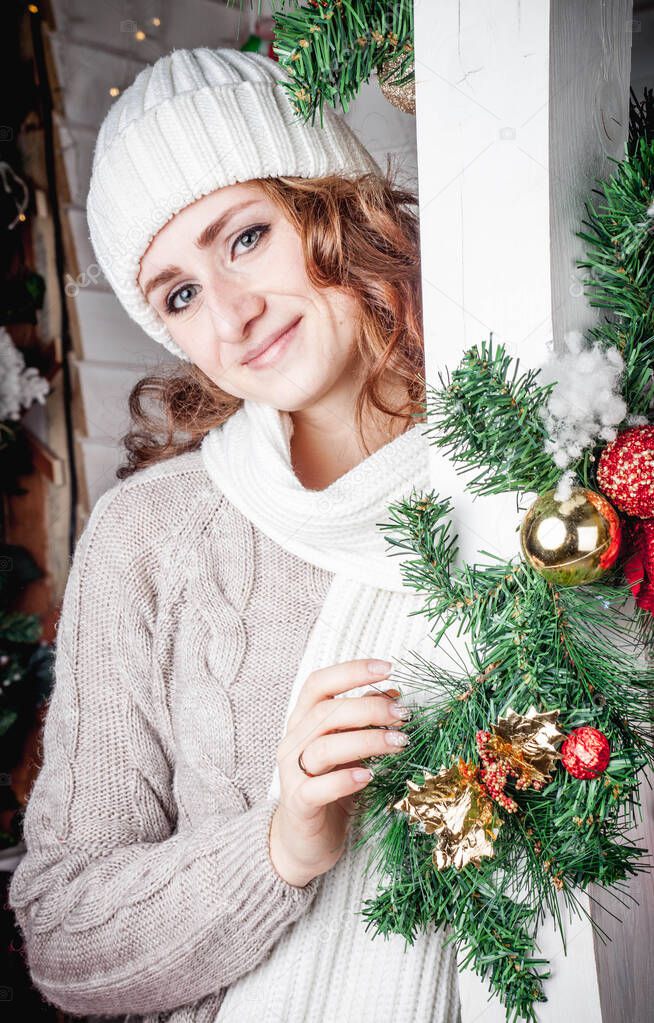 A red haired girl in a white knitted hat and a knitted sweater stands on a porch decorated in Christmas style. Winter