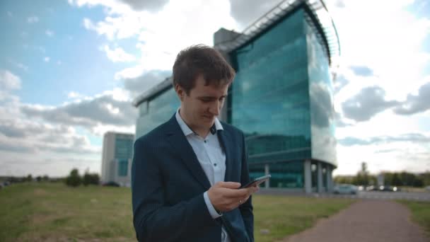 Gutaussehender Geschäftsmann scrollt und schaut aufs Smartphone. Hintergrund Bürogebäude — Stockvideo