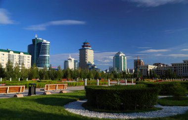 Parkın manzarası, beyaz pencere çerçeveli ahşap bankların yanında, yuvarlak yeşil bir çiçek tarlası, mavi gökyüzü ve birkaç bulut..