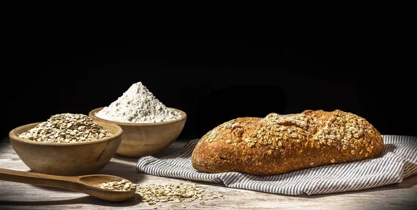 Bread still life with black background. Oatmeal bread with wooden bowl, linen sack, flour and oatmeal on rustic old wood