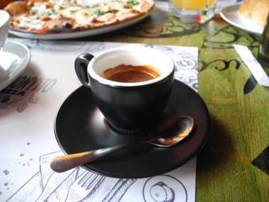 Masanın üzerinde küçük bir fincan sıcak espresso, küçük bir kaşık çay tabağında uzanıyor, arka planda pizza.