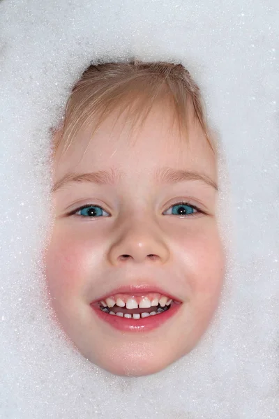 girl washes in the bathroom with white foam, above the water face with blue eyes and a smile with white teeth. Hygiene and body care is a healthy lifestyle.