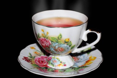 Siyah arka planda fincan tabağı olan beyaz porselen çay bardağı. Çiçeklerle boyanmış bir fincan. Çaydan beyaz buhar yükseliyor..