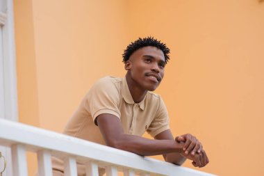 Genç adam balkonda dikiliyor ve önüne bakıyor.