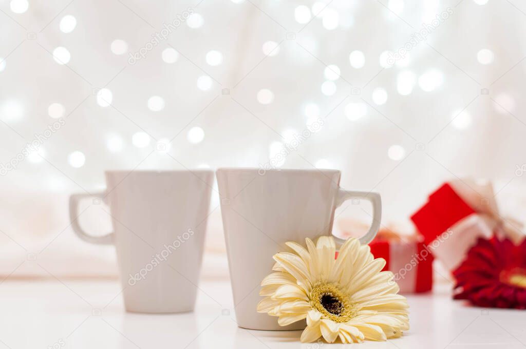 red gift boxes with ribbons, a pair of white cups and flowers on a white delicate background with bokeh.