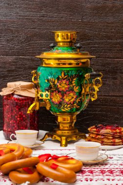 Karanlık bir arkaplanda dikey fotoğraf. Geleneksel desenli bakır semaver. Kırmızı kurdeleli simitler semaverde ve kreplerin yanında asılı duruyor. Rus çay seremonisi. Masa örtüsünde iki fincan..