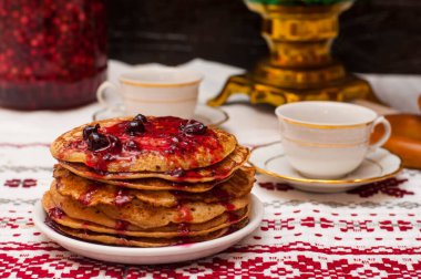 Karanlık bir arkaplanda yatay fotoğraf. Maslenitsa. Ön planda reçelli gözleme, bakır semaver. Rus çay seremonisi. Masa örtüsünde iki fincan..