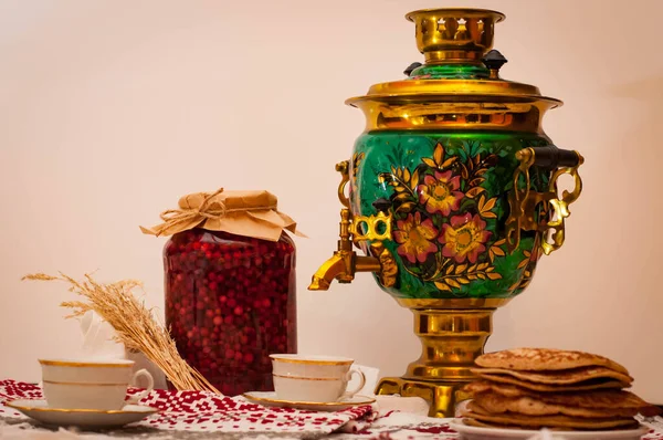 Foto horizontal sobre fondo blanco. samovar de cobre con un patrón tradicional. junto a panqueques y un tarro de mermelada. Ceremonia del té ruso. dos tazas en un mantel popular . —  Fotos de Stock