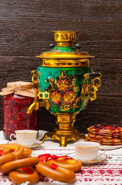 Vertical photo on a dark background. copper samovar with a traditional pattern. Bagels on a red ribbon hang on the samovar and next to pancakes. Russian tea ceremony. two cups on a folk tablecloth. — Stock Photo, Image