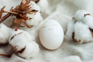 Yüzü olan beyaz Paskalya yumurtası. Yanında gri keten bir arka planda pamuktan bir dal var.