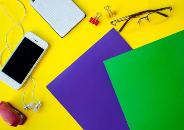 Sarı arka planda boş ekranlı telefon, beyaz elektrikli süpürge kulaklıklar, not defteri, ataçlar ve diğer ofis malzemeleri. Model yap. Kopyalama alanı olan pankart. İş konsepti ya da okula dönüş.