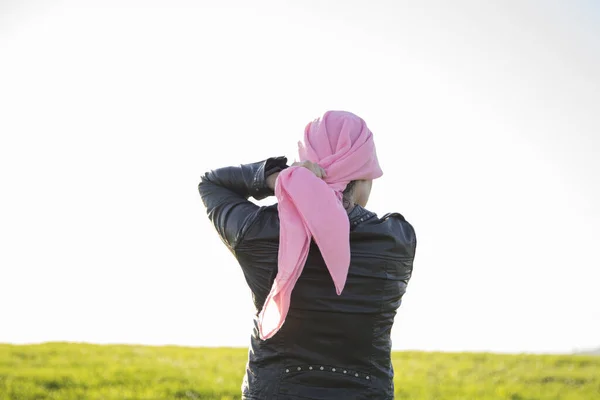 Young woman on her back in a black jacket and green pants wearing a pink headscarf in a green field. Concept of breast cancer.