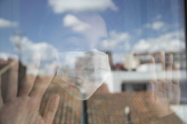 Maskeli ev hanımı kız pencereden dışarı bakıyor. Şehir ve bulutlar cama yansıyor. Pencerede iki eliyle sokağa geri dönmek için can atıyor. Konsept covid-19.