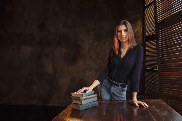 A charming European girl stands at the table and holds her hand on books. The background is dark and lit by window light.