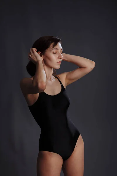 A young girl in a black swimsuit straightens her hair. The action takes place on a gray background. — Stock Photo, Image