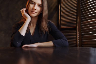 Esmer, ahşap masada oturuyor ve çerçeveye bakıyor. Işık huzmeleri pencereden içeri giriyor.