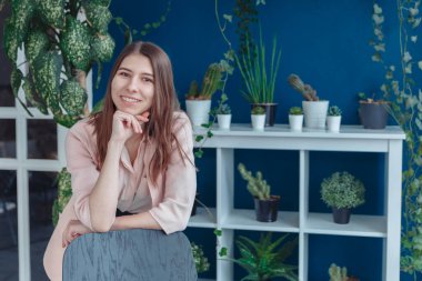 Gömlek giymiş gülümseyen bir kız çerçeveye bakıyor. Arka planda, çok sayıda kapalı alan bitkisi var. Pembe gömlekli gülümseyen bir kız çerçeveye bakıyor. Duvar maviye boyanmış.