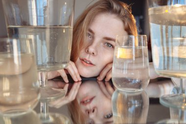A blonde with cute features looks into the frame. The girl is surrounded by water containers.