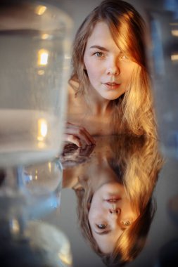 Kız doğrudan çerçeveye bakıyor. Kız aşağıdaki aynaya yansıyor. Kızın üzerine parlak bir ışık hüzmesi düşer. Işığın sıcak, güneşli bir tonu var. Kızın etrafında su dolu tanklar var..