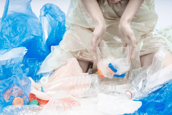 Elleri su ve plastikle dolu büyük bir bardağı tutuyor. Etrafta bir sürü tek kullanımlık tabak ve plastik var..