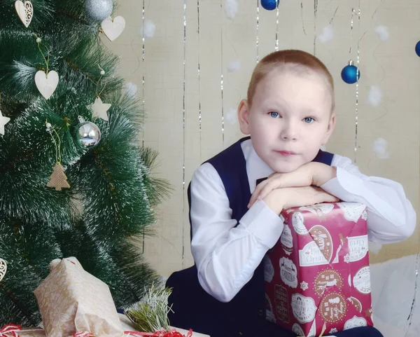 Beyaz gömlekli ve mavi yelekli bir çocuk Noel ağacında hediyelerle birlikte. Yeni yıl ve Noel tatili. Çocukların neşesi.