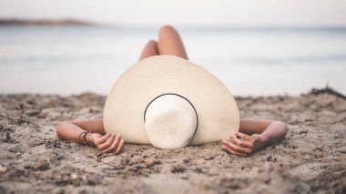 Genç bir kadın, Mallorca sahilinde, kumsalda, güzel, krem renkli şapkasıyla denizi seyrediyor. Kadın dinleniyor güneşli yaz tatillerinde Güzel havanın tadını çıkarıyor.