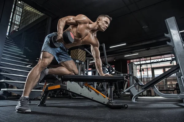 Gespierde jonge man tillen gewichten in de sportschool op donkere achtergrond. — Stockfoto