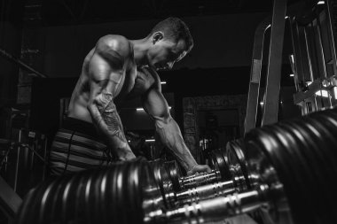 Kas yığını atletik vücut geliştirici adam spor salonunda koyu arka planda, halterli dramatik ışık altında sıkı çalışıyor.
