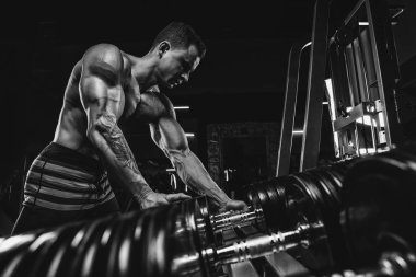 Kas yığını atletik vücut geliştirici adam spor salonunda koyu arka planda, halterli dramatik ışık altında sıkı çalışıyor.