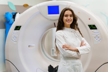Tıp önlüklü genç kadın doktor kameraya bakıyor. Bilgisayar tomografisi üstüne hasta. Tıp kliniğinde CT taraması