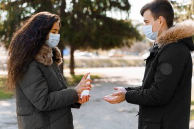 Tıbbi maskeli ve antiseptik spreyli genç bir çift. Coronavirüs kovid 19 'u durdurmak için erkek arkadaşının ellerini spreyliyor.