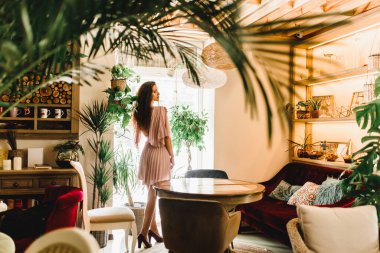 Profesyonel model pencerenin karşısında duruyor ve sağ tarafa bakıyor. Uzun dalgalı saçlı doğal güzel kadınlar. Büyük yeşil bitkileri ve palmiyeleri olan bir kafe.