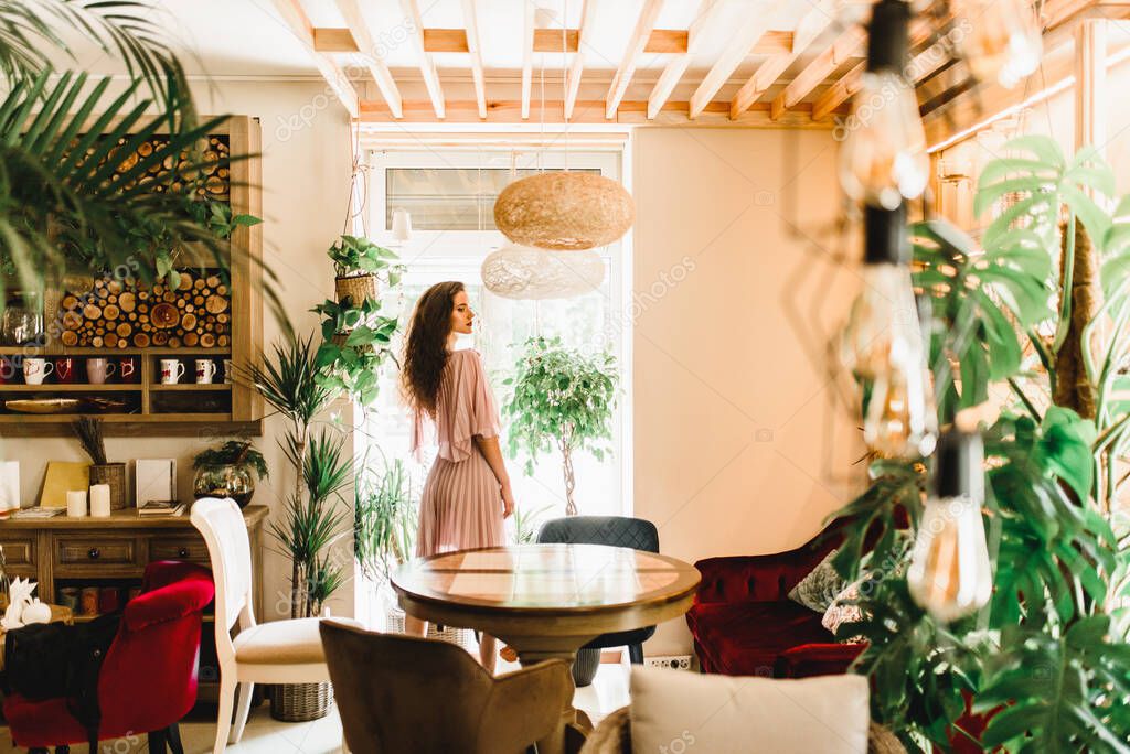Professional model is standing opposite to window and looking to the right side. Natural beauty women with long wavy hair. Cafe with big green plants and palm tree