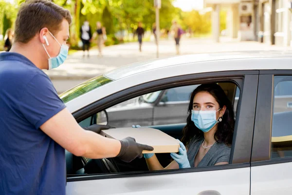 Safe food delivery from pizzeria to car during quarantine coronavirus. Attractive business woman in medical mask and gloves gets pizza in cardboard box. Delivering by car according social distance