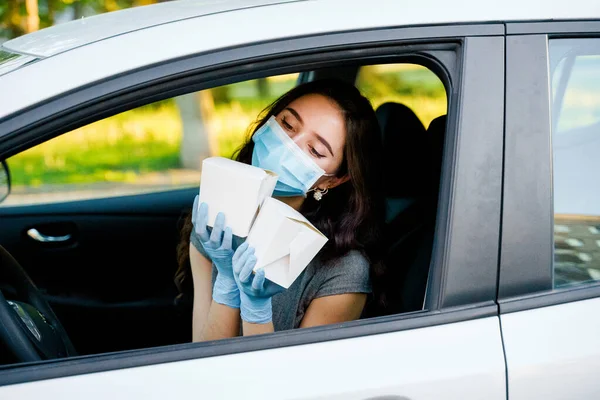 Young attractive girl in car in medical gloves and mask holds wok in box udon noodles with tempuru, shrimps, in hands and smiles. Udon noodles in white box delivery. 2 Wok box udon advertise 1+1.