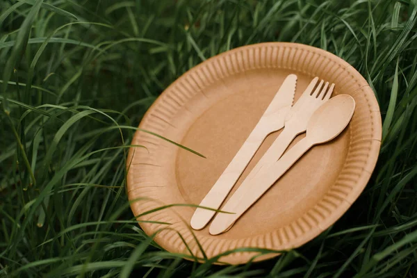 Eco-friendly natural plate with spoon, fork, knife. Set of disposable ecological dishes on green grass background. Sustainability of planet. Cardboard dishes made of fiber of bamboo and bagasse