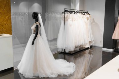 İpek şifon, tül ve dantelden yapılmış gelinlik giymiş aptal. Düğün dükkanının içinde. Düğün salonundaki askılarda beyaz krem gelinlik. Kollarında inciler ve kristaller var..