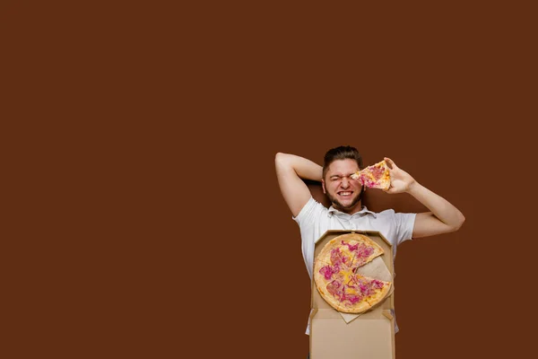 レストランからオンラインピザの安全配達サービス 若い幸せな宅配便の男は茶色の背景に横たわり ピザで周りの愚か者 広告のための空きスペース チーズボード付きピザ — ストック写真