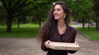 Pizzacıdan yemek servisi. Kurye kız pizza kutularıyla yeşil parkta yürüyor ve gülümsüyor. Pizza teslimatı için yönlendir. Güvenli fast food teslimatı. Evde kal.