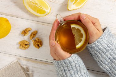 Sıcak süveter giymiş, beyaz ahşap masada limonlu çay tutan kadın elleri. Alternatif tıp konsepti. Güvenli tedavi. Üst görünüm