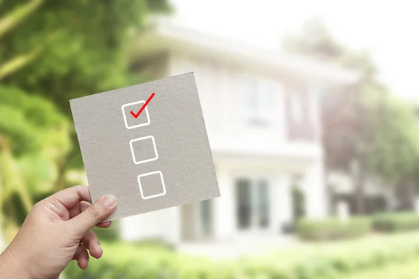 Hand hält braunes Papier mit Häkchensymbol gegen neues Haus — Stockfoto