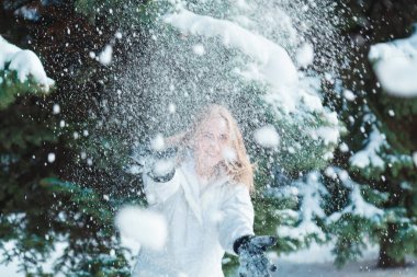 Soğuk bir kış günü genç güzel neşeli kadın beyaz ceketli uzun sarı saçlı kar ağaçlar karla kaplı zemin karşı oynadığı ve neşeyle gülüyor