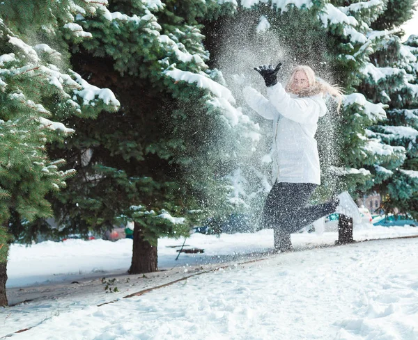 У холодний зимовий день молода красива весела жінка з довгим світлим волоссям у білій куртці грає зі снігом на тлі засніжених дерев і весело сміється — стокове фото