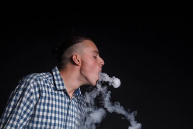 Mavi ekose gömlekli şık bir saç kesimi olan genç bir adam siyah bir zemin üzerine bir sigara elektronnuju içiyor. Egzoz sigara yapar duman halkaları rakamlar